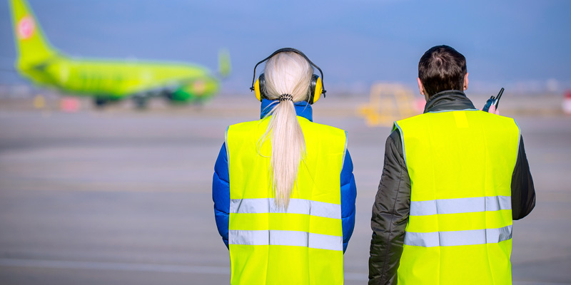 Två flygplatstekniker i reflexvästar som står på plattan och blickar mot ett parkerat flygplan