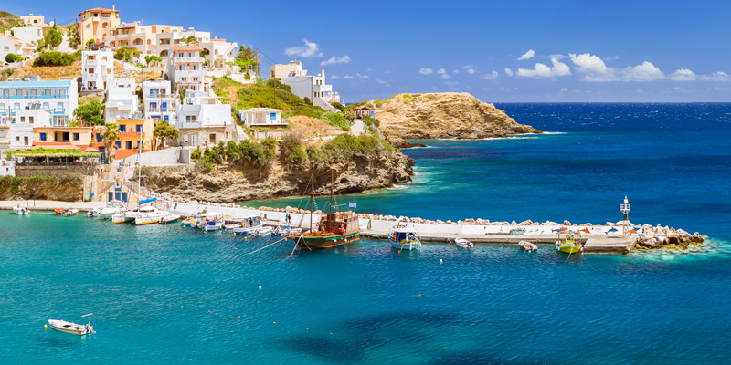 Pittoresk hamn på Kreta med vita hus längs klippan, småbåtar och klart blått vatten